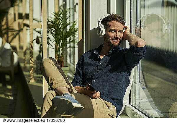 Smiling male freelancer with eyes closed listening music through wireless headphones by window
