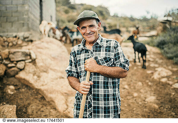 Smiling male goat herder holding broom while standing at farm