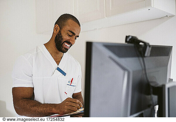 Smiling male healthcare worker writing while consulting patient through video call