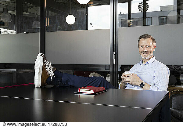 Smiling male professional with feet up having coffee while sitting at table tennis table in office