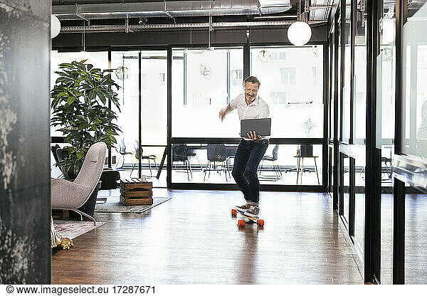 Smiling male professional with laptop riding skateboard at office