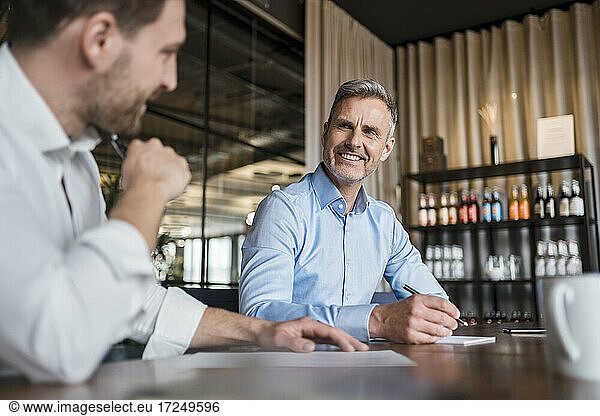 Smiling male professional with pen discussing with colleague at work place