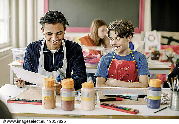 Smiling male student showing painting to teenage friend during art class at high school