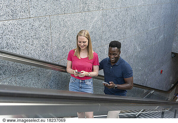 Smiling man and woman using smart phones moving up on escalator by wall