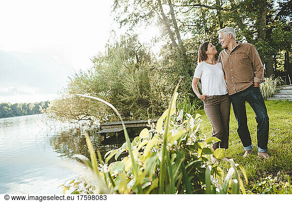 Smiling man and woman with hands in pockets looking at each other by lake
