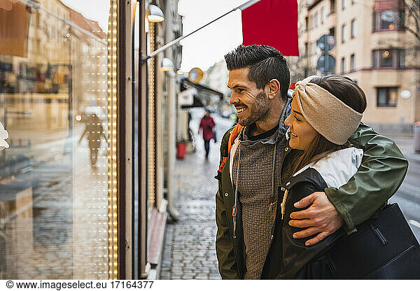 Smiling man arm around woman while doing window shopping in city