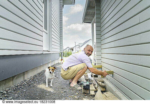 Smiling man building wooden wall crouching by dog near house