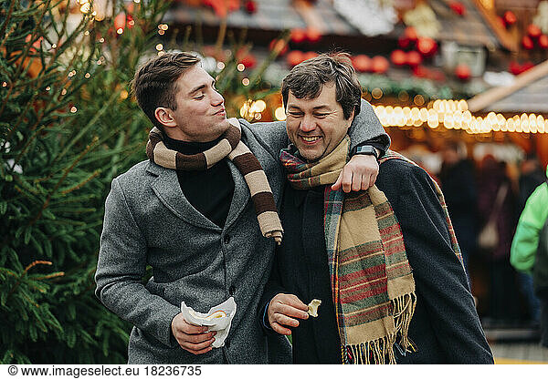 Smiling man eating trdelnik with arm around father enjoying at Christmas market