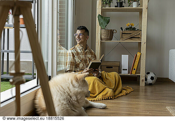 Smiling man holding book looking out of window at home