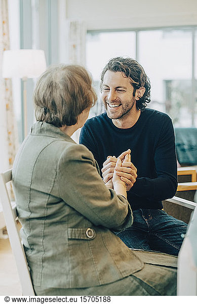 Smiling man holding hands while talking to grandmother at elderly nursing home