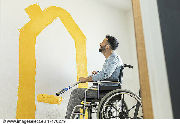 Smiling man holding paint roller while sitting on wheelchair in living room