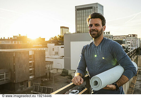 Smiling man holding rolled up exercise mat leaning on railing at terrace