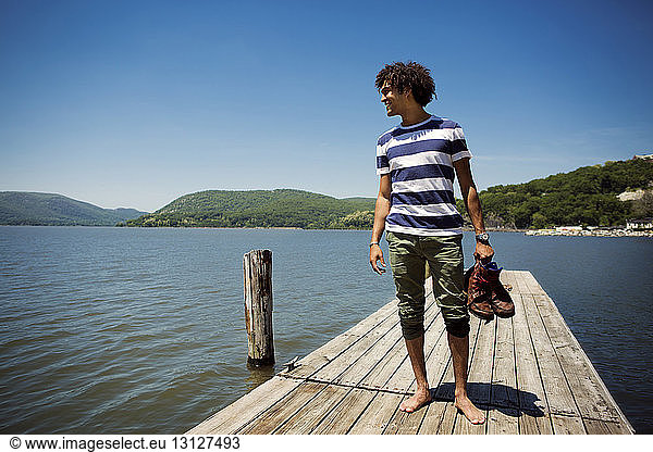 Smiling man holding shoes while standing on jetty over lake