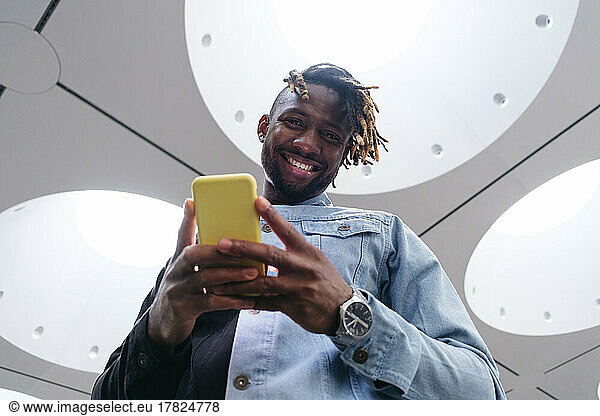 Smiling man holding smart phone standing under illuminated ceiling
