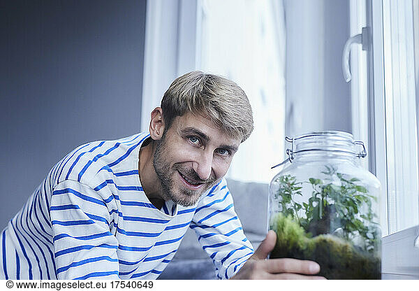 Smiling man leaning by plant jar near window at home