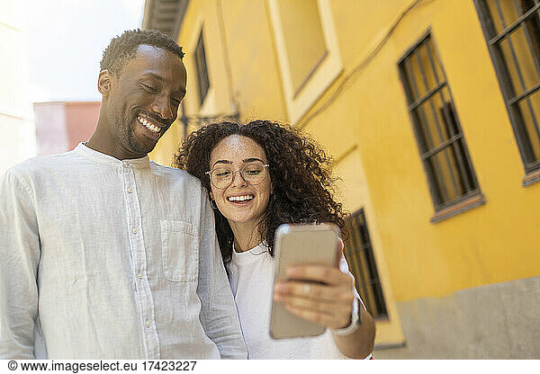 Smiling man looking at smart phone held by woman near building