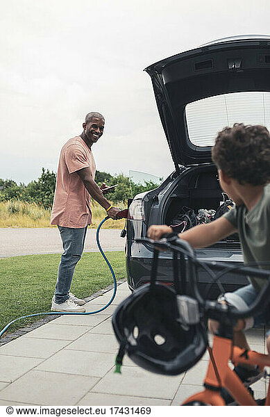 Smiling man looking at son riding bicycle while charging electric car