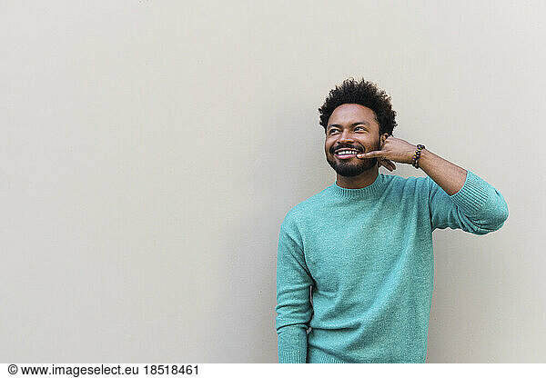Smiling man making calling gesture in front of wall