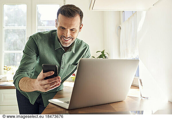 Smiling man making online payment with mobile phone and laptop in the kitchen