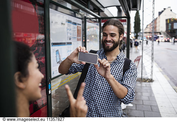 Smiling man photographing female friend through smart phone at bus stop in city
