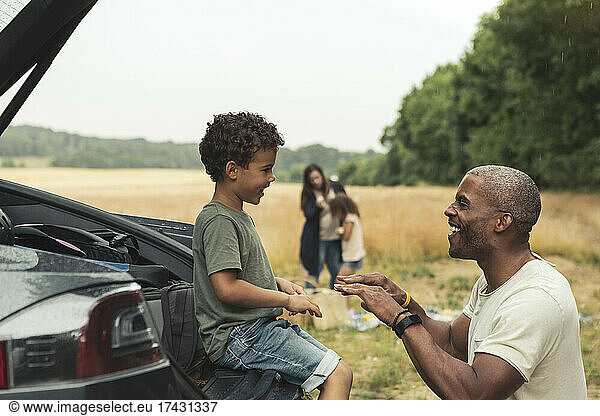 Smiling man playing with son sitting in car trunk during picnic