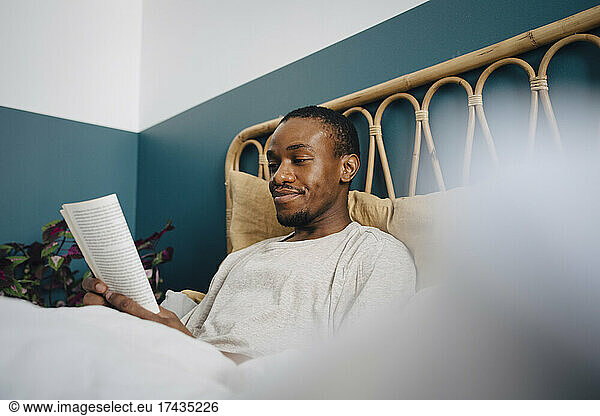 Smiling man reading book while sitting on bed at home