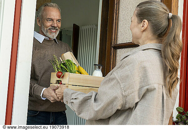 Smiling man receiving crate of groceries from woman at door