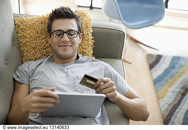 Smiling man shopping online on tablet while lying on sofa at home