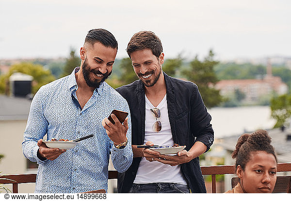 Smiling man showing mobile phone to friend while holding meal during party on terrace