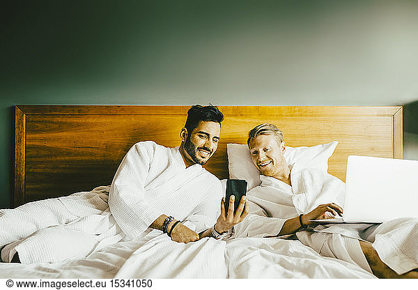 Smiling man showing smart phone to boyfriend using laptop while leaning in bed at hotel