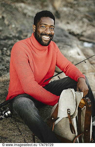 Smiling man sitting with backpack and fishing rod on rock