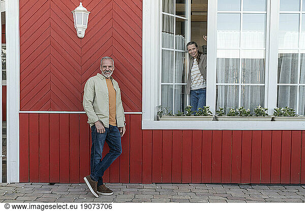 Smiling man standing by woman looking from house window