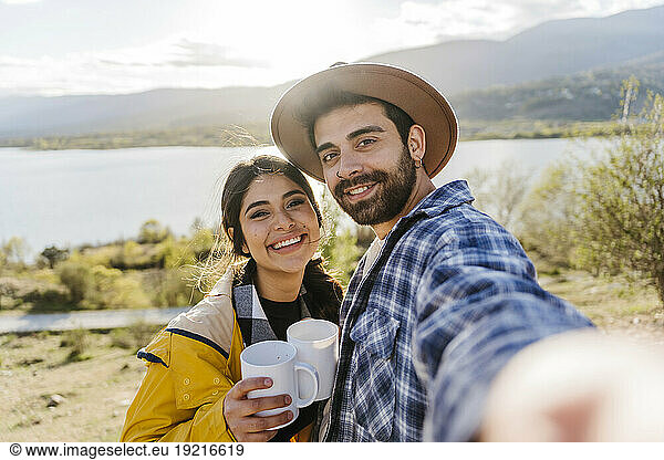 Smiling man taking selfie with girlfriend holding coffee mug