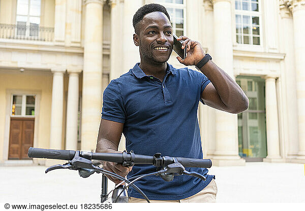 Smiling man talking on phone standing with bicycle in front of building
