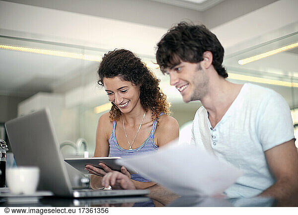 Smiling man using laptop while holding document by girlfriend in kitchen