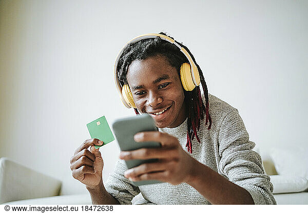 Smiling man using mobile phone with credit card at home