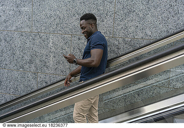 Smiling man using smart phone moving down on escalator