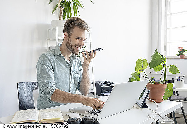 Smiling man using smartphone and laptop at desk in office