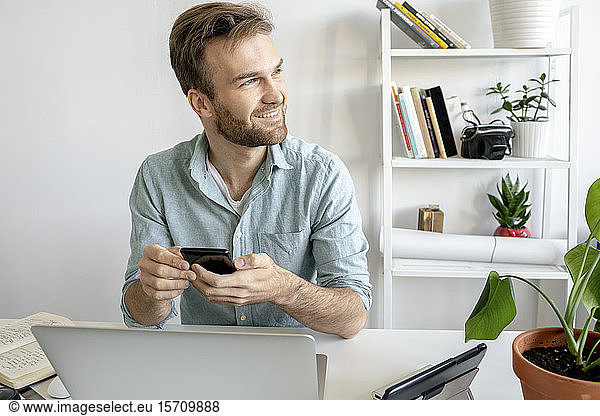 Smiling man using smartphone at desk in office looking sideways