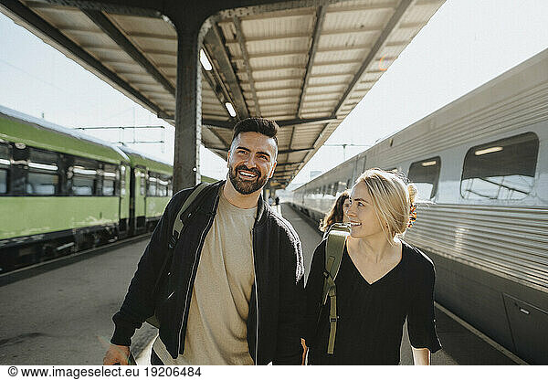 Smiling man walking with woman near train at railroad station