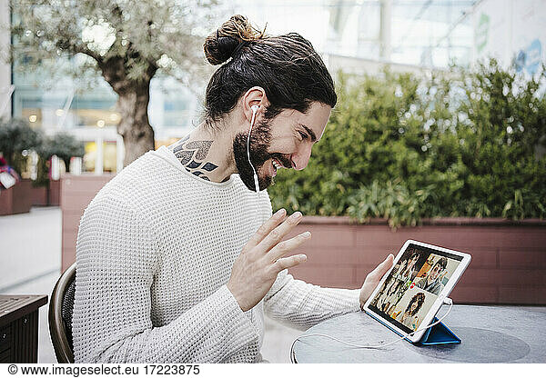 Smiling man waving hand during video call through digital tablet at sidewalk cafe