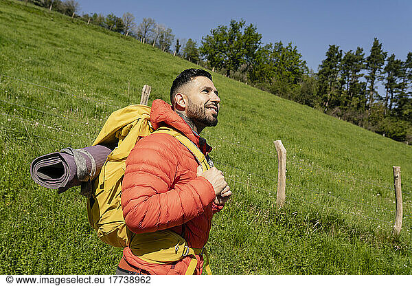Smiling man wearing backpack standing in front of meadow on sunny day
