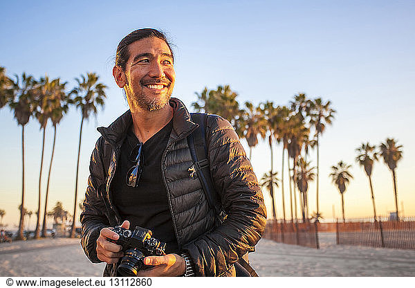 Smiling man wearing jacket while holding camera at beach