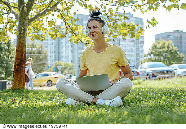 Smiling man wearing wireless headphones sitting with laptop in grass