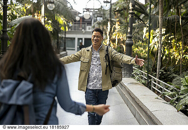 Smiling man with arms outstretched walking towards woman on footpath in park