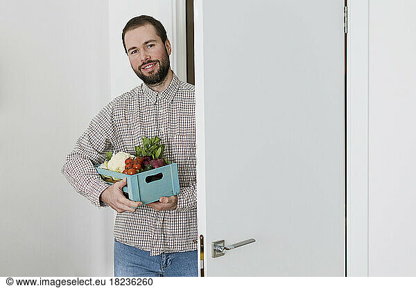 Smiling man with crate of fresh vegetables standing at doorway