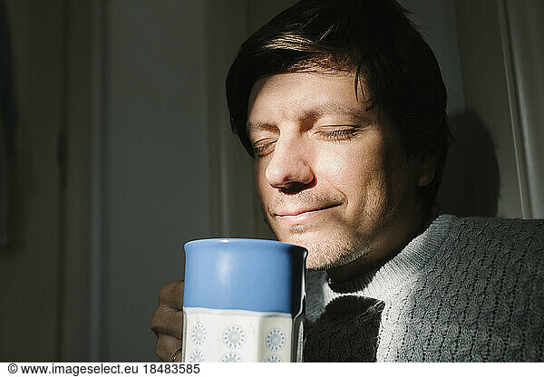 Smiling man with eyes closed holding cup of tea at home