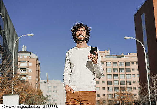 Smiling man with eyes closed holding smart phone under blue sky