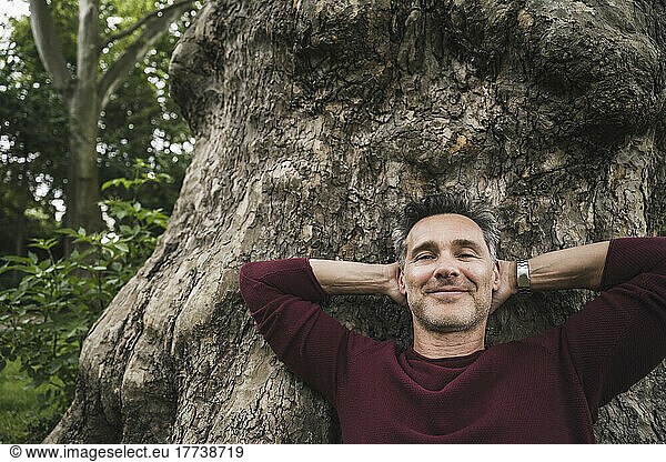 Smiling man with hands behind head leaning on tree trunk