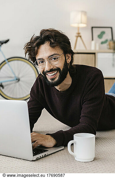 Smiling man with laptop and coffee cup lying on carpet at home